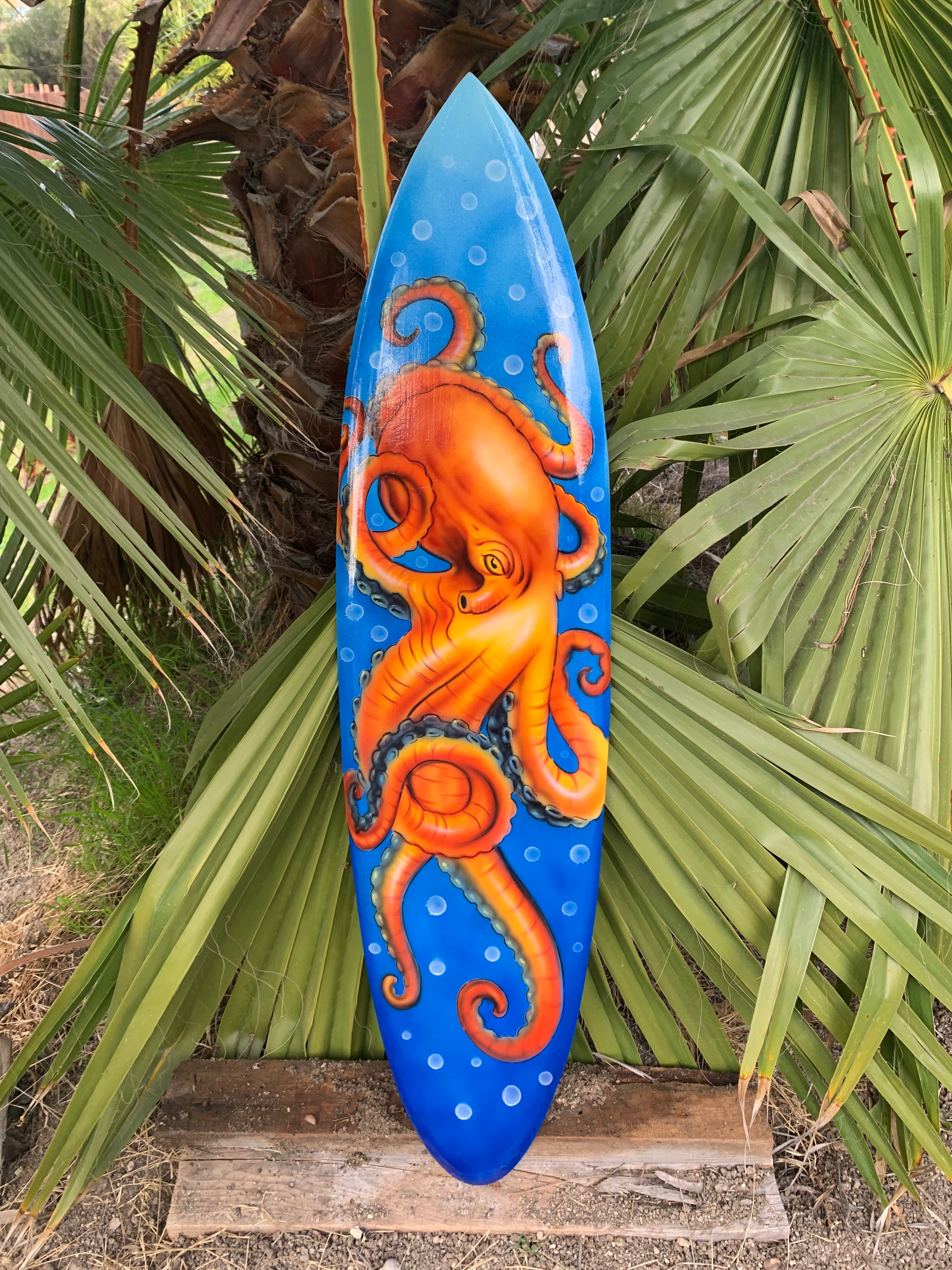 Blue surfboard with bright orange octopus design leaning against palm leaves